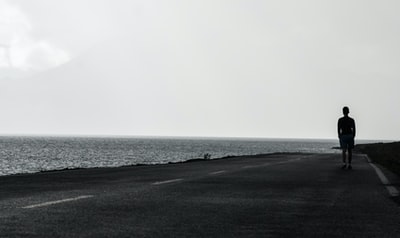 男人站在灰色的天空下沥青道路旁边的水域
